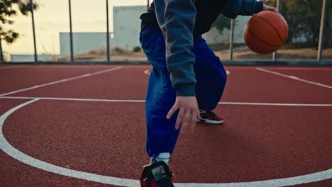 Primer-Plano-De-Una-Chica-Rubia-Con-Uniforme-Deportivo-Y-Pantalones-Azules-Maniobra-Y-Golpea-Una-Pelota-Naranja-Desde-El-Suelo-Rojo-En-Una-Cancha-De-Baloncesto-Callejera-Durante-Un-Partido-Deportivo-Por-La-Mañana.