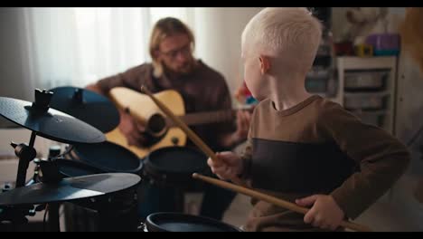 Un-Niño-Albino-Feliz-Con-Cabello-Blanco-Toca-Una-Batería-Electrónica-Especial-Usando-Baquetas-Amarillas-Y-Su-Padre,-Un-Hombre-Rubio-De-Cabello-Largo-Con-Barba-Y-Gafas,-Toca-La-Guitarra-Con-Su-Hijo.