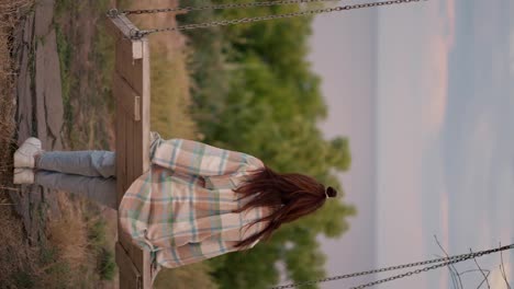 Vertical-video:-A-brunette-in-a-checkered-shirt-swings-on-a-wooden-swing-outside-the-city-on-the-weekend.-Rest-in-the-country-by-the-sea