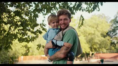 Happy-dad-with-curly-hair-and-Green-T-shirt-together-with-his-little-son-in-a-Blue-sweater-with-blond-hair-in-the-park-near-the-skatepark.-Father-holding-his-happy-little-son-in-his-arms