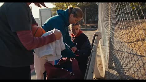 Three-blonde-girls-in-sportswear-put-their-things-in-sports-bags-after-training-on-an-outdoor-basketball-court-on-a-bench-near-a-lattice-fence-on-a-sunny-summer-day