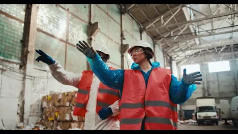 A-brunette-girl-in-safety-glasses-and-a-special-uniform-in-an-orange-vest-and-a-white-helmet-walks-with-her-employee-a-man-with-Black-skin-through-a-huge-waste-paper-processing-plant