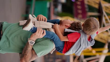 Vertical-video-of-a-father-in-a-Green-T-shirt-and-his-little-boy,-his-blond-son-with-blue-eyes-in-a-red-T-shirt-sits-on-his-shoulders-and-does-not-look-at-the-rides-in-an-amusement-park-during-the-day