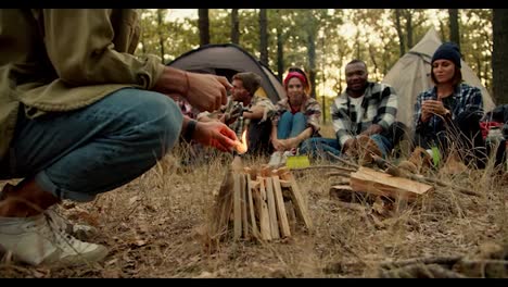 Close-up-shot:-a-man-in-a-light-green-jacket-makes-a-fire-in-the-forest-during-a-halt-with-the-rest-of-the-hikers-against-the-backdrop-of-tents-in-a-green-autumn-forest-and-withered-yellow-grass