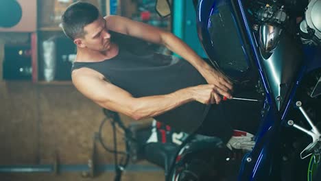 Vertical-video-of-a-man-with-a-short-haircut-in-a-gray-T-shirt-unscrewing-bolts-on-his-motorcycle-for-repairs-and-repairs-in-his-garage-workshop