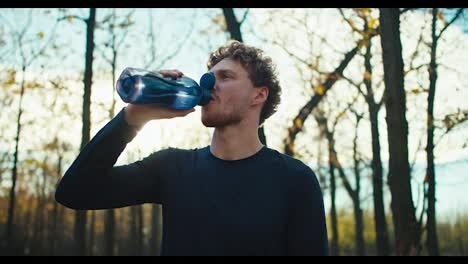 A-happy-man-with-curly-hair-and-a-beard-in-a-black-sports-uniform-drinks-water-from-a-gray-sports-bottle-and-pours-himself-over-it-to-relax-after-jogging-in-the-morning-in-autumn