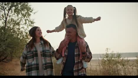 A-little-blonde-girl-is-having-fun-while-sitting-on-the-shoulders-of-her-brunette-dad-in-a-brown-checkered-shirt-who-is-running-with-his-wife-while-walking-along-a-path-outside-the-city