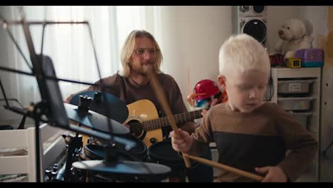 Un-Niño-Albino-De-Pelo-Blanco-Con-Un-Corte-De-Pelo-Corto-Y-Una-Chaqueta-Marrón-Toca-Una-Batería-Electrónica-Usando-Baquetas-Especiales-Junto-Con-Su-Padre,-Un-Hombre-Rubio-Con-Gafas-Y-Barba-Que-Toca-Una-Guitarra-Acústica-Con-Su-Hijo.