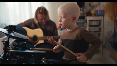 Happy-albino-boy-with-white-hair-energetically-plays-the-drum-set-with-the-help-of-special-sticks-and-his-dad,-a-blond-man-with-a-beard-and-glasses,-plays-an-acoustic-guitar-with-his-son-in-a-room-for-developing-talents-for-playing-musical-instruments-at-home