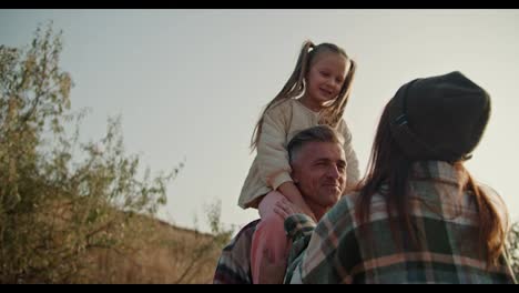 A-middle-aged-brunette-man-with-a-little-gray-hair-communicates-with-his-wife-in-a-hat-and-a-plaid-shirt-while-his-little-daughter-sits-on-his-shoulders-during-his-picnic-outside-the-city-in-the-summer