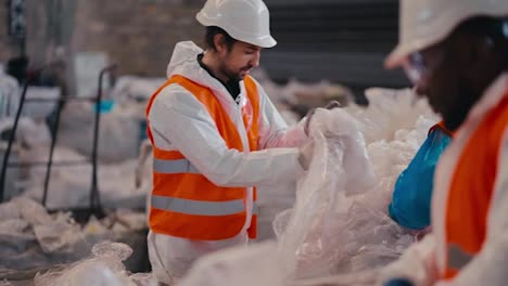 A-brunette-man-with-a-beard-in-a-white-protective-uniform-and-an-orange-vest-presses-cellophane-and-polyethylene-together-with-his-colleagues-at-a-large-waste-recycling-plant