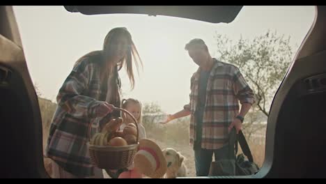 A-middle-aged-man-with-gray-hair-in-a-plaid-shirt-and-his-wife,-a-brunette-girl-in-a-green-plaid-shirt,-together-with-their-daughter,-are-packing-things-that-are-necessary-for-a-picnic-while-driving-back-home-after-a-walk-in-the-summer