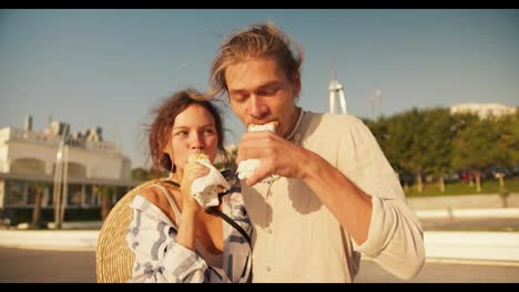 Happy-couple-portrait-guy-and-girl-eating-hot-dog-on-modern-beach-in-the-morning.-Blond-guy-in-light-clothes-III-brown-haired-girl-goes-hot-dogs-on-a-sunny-beach-in-the-morning