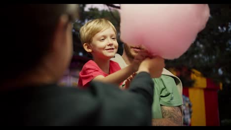 A-little-blond-boy-in-a-red-T-shirt-takes-a-large-cotton-candy-from-the-hands-of-a-cashier-in-an-amusement-park-while-sitting-in-the-arms-of-his-father-in-a-Green-T-shirt