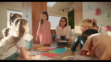 A-female-teacher-with-a-bob-hairstyle-in-glasses-and-a-white-shirt-conducts-a-drawing-lesson-for-preschool-children.-A-group-sits-on-the-floor-and-draws-on-multi-colored-paper-using-pencils-in-a-special-room-in-a-club-for-preparing-children-for-school