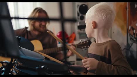 Primer-Plano-De-Un-Niño-Albino-Feliz-Con-Cabello-Blanco-Y-Un-Corte-De-Pelo-Corto-Tocando-Batería-Electrónica-Usando-Baquetas-Especiales-Para-Instrumentos-De-Percusión-Y-Su-Padre-Tocando-Una-Guitarra-Acústica-Con-él,-Un-Hombre-Rubio-Feliz-Con-Barba-Y-Gafas-En-La-Habitación