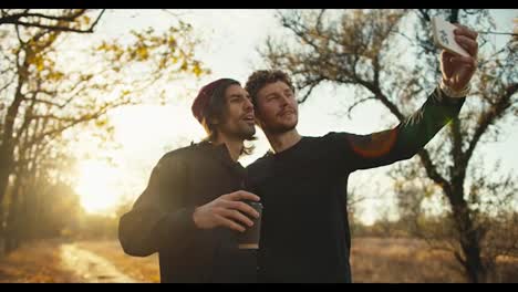 Happy-man-with-curly-hair-in-a-black-sports-uniform-takes-a-selfie-with-his-male-friend-in-a-red-cap-with-a-cardboard-cup-of-coffee-near-an-autumn-forest-with-brown-foliage-in-the-morning-at-sunrise