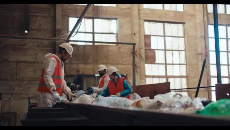 Ein-Team-Aus-Drei-Recyclinganlagenarbeitern-In-Weißen-Uniformen-Und-Orangefarbenen-Westen-Ordnet-Flaschen-In-Einer-Großen-Alten-Recyclinganlage-Nach-Der-Farbe-Des-Kunststoffs