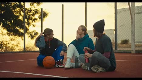 A-blonde-girl-in-a-sports-uniform-is-warming-up-and,-together-with-her-friends,-sits-on-the-red-floor-on-a-street-basketball-court-and-communicates-with-them-at-sunrise