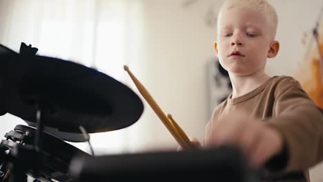 Close-up-shot-from-below-of-a-happy-blond-albino-boy-with-white-hair-setting-up-an-electronic-drum-kit-before-starting-to-play-it-with-wooden-sticks-in-his-room,-developing-his-talent-and-hobby