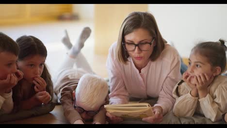 Close-up-shot-of-a-blonde-woman-with-glasses-in-a-pink-shirt-reading-a-book-to-children-in-a-club-for-preparing-children-for-school-in-a-special-room-Lying-on-pillows-on-the-floor