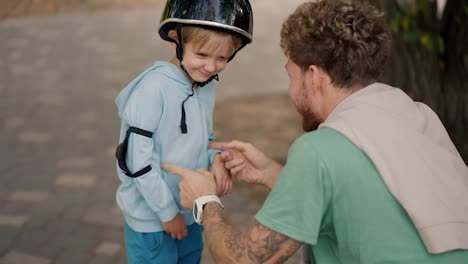 Un-Niño-Rubio-Con-Un-Suéter-Azul-No-Quiere-Usar-Un-Casco-Protector-Negro-Para-Andar-En-Patineta-En-Un-Parque-De-Patinaje,-Que-Su-Padre-Con-Cabello-Rizado-Y-Una-Camiseta-Verde-Le-Puso.