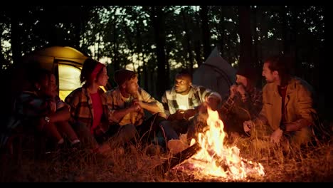 Eine-Gruppe-Von-Pfadfindern-Erzählt-Sich-Während-Einer-Rast-Gegenseitig-Gruselgeschichten-In-Der-Nähe-Eines-Feuers-Im-Nächtlichen-Wald-Mit-Zelten-Im-Hintergrund