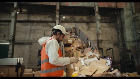 A-brunette-man-with-a-beard-in-a-white-helmet-and-a-white-uniform-in-an-orange-vest-stands-near-a-conveyor-belt-at-a-waste-paper-recycling-plant