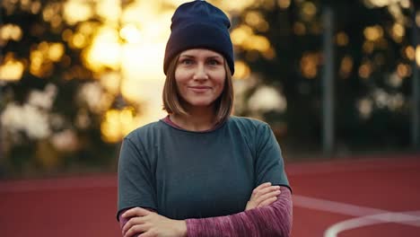 Close-up-portrait-of-a-confident-and-happy-blonde-girl-in-a-black-hat-and-in-a-sports-uniform-who-folds-her-arms-on-her-chest-and-looks-at-the-camera-in-the-morning-at-Sunrise