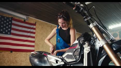 A-brunette-mechanic-girl-in-a-black-top-and-blue-overalls-repairs-a-motorcycle-in-her-workshop-against-the-backdrop-of-the-USA-flag.-Working-as-a-mechanic,-girl-repairing-a-motorcycle