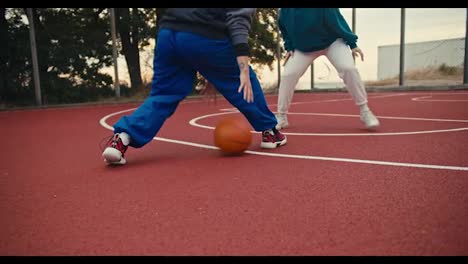 Toma-De-Primer-Plano-De-Una-Niña-Con-Pantalones-Azules-Maniobrando-Y-Golpeando-La-Pelota-Desde-El-Suelo-Mientras-Juega-Baloncesto-Con-Una-Niña-Con-Pantalones-Blancos-En-Una-Cancha-De-Baloncesto-Roja-Temprano-En-La-Mañana.