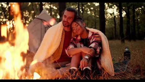 A-brunette-guy-and-a-Black-skinned-girl-are-sitting-and-hugging-under-a-white-blanket-near-a-fire-against-the-backdrop-of-a-tent-in-a-green-forest-during-a-hike.-Happy-and-cozy-evening-during-a-rest-in-the-evening-during-a-hike