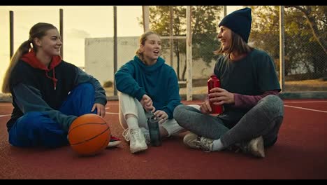 Close-up-a-trio-of-blonde-girls-in-sports-uniforms-hold-sports-bottles-in-their-hands,-sit-on-the-red-floor-on-a-street-court-near-an-orange-basketball-ball,-communicate-and-warm-up-in-the-morning