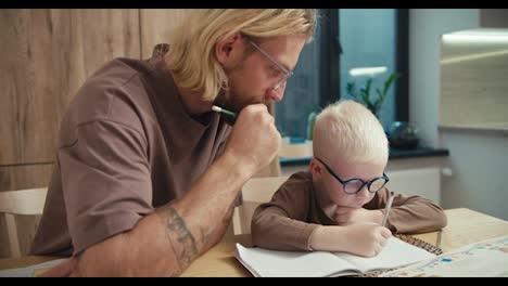 Ein-Kleiner-Albinojunge-Mit-Weißen-Haaren-Und-Blauer-Brille-Hält-Einen-Stift-In-Der-Hand-Und-Schreibt-Etwas-In-Sein-Notizbuch.-Sein-Vater,-Ein-Blonder-Mann,-Hilft-Ihm-Bei-Den-Hausaufgaben