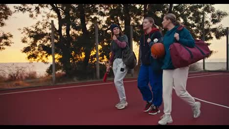 Side-view-of-a-happy-blonde-girl-in-a-turquoise-hoodie-walking-and-carrying-a-basketball-while-chatting-with-her-two-friends-after-a-basketball-match-in-the-evening-at-sunset