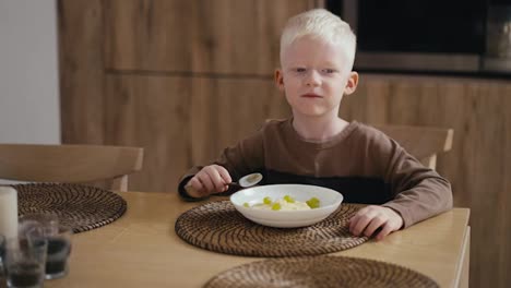 Happy-little-albino-boy-with-white-hair-color-and-blue-eyes-eats-oatmeal-with-green-grapes-while-sitting-on-a-modern-chair-in-the-kitchen.-Morning-before-school-day