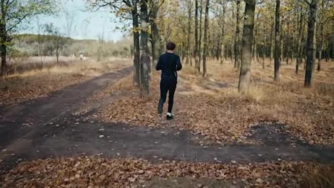 A-brunette-man-with-curly-hair-in-a-black-sports-uniform-runs-through-the-autumn-forest-along-an-earthen-path-and-looks-at-his-watch-in-the-morning