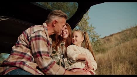 Close-up-shot-of-a-happy-middle-aged-brunette-man-with-a-little-gray-hair-talking-and-hugging-his-wife,-the-brunette-girl-in-a-green-checkered-shirt,-and-a-little-blonde-daughter-Sitting-in-the-open-trunk-of-a-car-on-his-picnic-Far-from-the-city