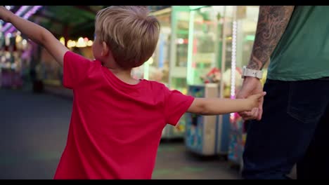 Ein-Glücklicher-Kleiner-Blonder-Sohn-In-Einem-Roten-T-Shirt-Geht-Mit-Seinem-Vater-Los-Und-Schaut-Sich-Die-Fahrgeschäfte-Im-Vergnügungspark-An.-Ein-Kleiner-Junge-In-Einem-Roten-T-Shirt-Und-Sein-Vater-Schauen-Sich-Aktiv-Die-Fahrgeschäfte-Im-Vergnügungspark-An,-Während-Sie-An-Ihnen-Entlanggehen