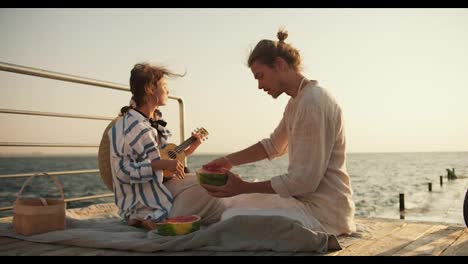 Ein-Mann-Und-Ein-Mädchen-Sitzen-Auf-Dem-Pier,-Ein-Mann-In-Leichter-Kleidung-Schneidet-Eine-Wassermelone-Auf,-Ein-Mädchen-Spielt-Ukulele-Und-Sie-Schauen-Auf-Das-Meer.-Ein-Mädchen-In-Einem-Blau-weißen-Hemd-Spielt-Und-Singt-Auf-Einem-Musikinstrument-Und-Ein-Mann-Schneidet-Dabei-Eine-Wassermelone-Für-Ein-Picknick-Auf-Dem-Pier-In-Der-Nähe-Des-Meeres.