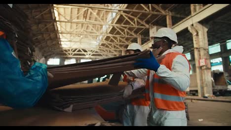 Ein-Trio-Von-Menschen-In-Weißen-Schutzuniformen-Und-Orangefarbenen-Westen-Sortiert-Riesige-Stapel-Altpapier-In-Einer-Abfallrecyclinganlage-Und-Lagert-Sie.-Zwei-Männer-Nehmen-Riesige-Pappbögen-Zusammen-Und-Bewegen-Sie