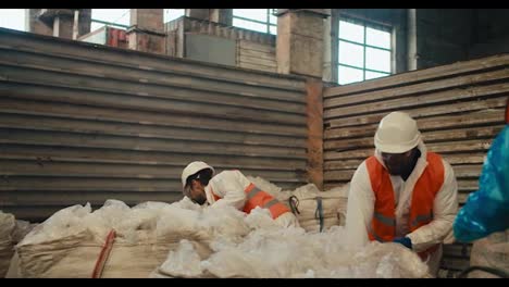 A-man-with-Black-skin-in-a-white-helmet-and-a-white-protective-uniform-in-an-orange-vest-walks-on-a-tramp-and-presses-cellophane-garbage-along-with-his-colleagues-a-man-with-a-beard-and-a-girl-in-a-blue-protective-suit