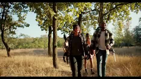 Front-view-of-a-happy-group-of-several-pairs-of-people-in-special-hiking-clothes-with-hiking-backpacks-walking-through-the-forest-near-dried-yellow-summer-grass.-A-Black-man-talks-to-a-blonde-girl-with-a-bob-hairstyle