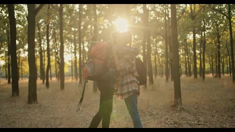 A-happy-blonde-girl-with-a-bob-hairstyle-in-a-plaid-shirt-with-a-large-red-hiking-backpack-and-a-brunette-girl-with-a-red-bandana-meet-during-a-hike-and-hug-against-the-backdrop-of-a-summer-sunny-forest