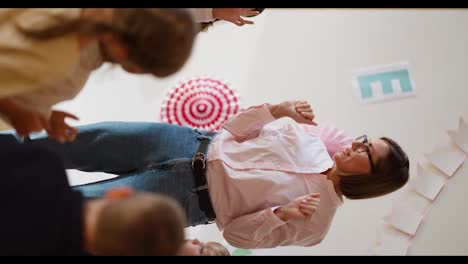 Vertical-video-of-a-woman-with-a-bob-hairstyle-in-glasses-in-a-pink-shirt-doing-a-warm-up-for-preschool-children-in-a-special-club-for-preparing-children-for-school