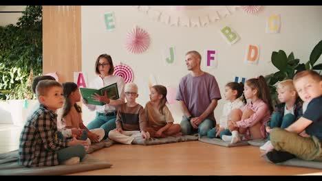 blonde-girl-in-glasses-with-a-bob-hairstyle-reads-a-green-book-for-preschool.-Children-children-sit-in-a-circle-and-listen-carefully-to-their-teacher.-The-second-teacher,-a-man-in-a-purple-T-shirt,-supports-and-listens-attentively-to-the-teacher