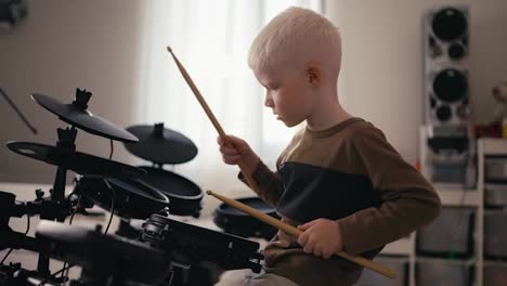 Shot-from-the-side-of-a-small-albino-boy-with-white-hair-and-blue-eyes-playing-and-tuning-his-electric-musical-drum-kit-using-special-wooden-sticks-during-his-rest-and-day-off-in-his-room