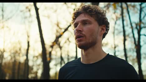 A-confident-and-serious-man-with-curly-hair-and-a-beard-in-a-black-sports-uniform-wipes-sweat-from-his-forehead-with-a-gray-towel-after-an-evening-jog-in-the-autumn-forest