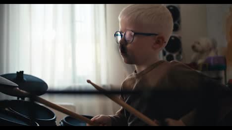 Close-up-shot-of-a-blond-albino-boy-with-glasses-playing-an-electronic-drum-set-using-special-sticks-and-listening-to-music-in-his-room.-Happy-little-boy-pursuing-his-hobby-and-developing-his-talent-of-playing-musical-instruments-at-home-in-his-room