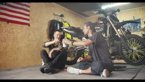 Two-happy-male-mechanics-drink-drinks-from-small-bottles-in-their-garage-workshop.-Break-and-rest-while-working-in-the-workshop.-Auto-mechanic-duet